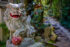 Color photo. A grey stone statue of a lion, in Japanese style, sitting on a pedestal. Its left front paw rests on a ball. The ball has a red-painted flower motif. The lion's claws, teeth, and eyes are painted white. Its mouth is painted red. Its pupils are painted gold.