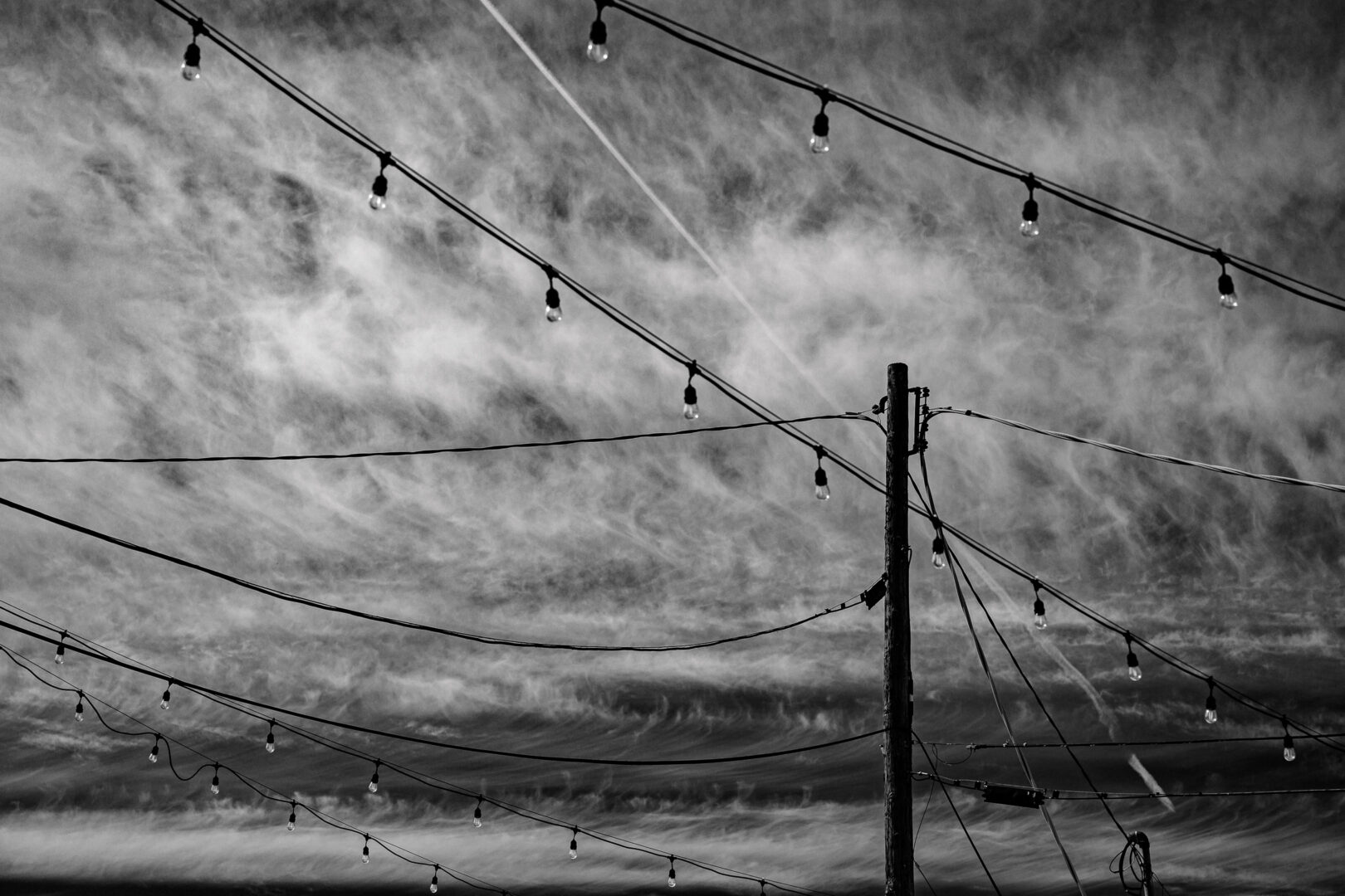 Black and white photo. A utility pole juts up from the bottom of the frame. Dark wires are attached to it, and cross from left to right. Other wires cross the frame diagonally from upper left to lower right, and these have lightbulbs hanging from them. Behind the pole and wires a sky full of wispy clouds is visible.