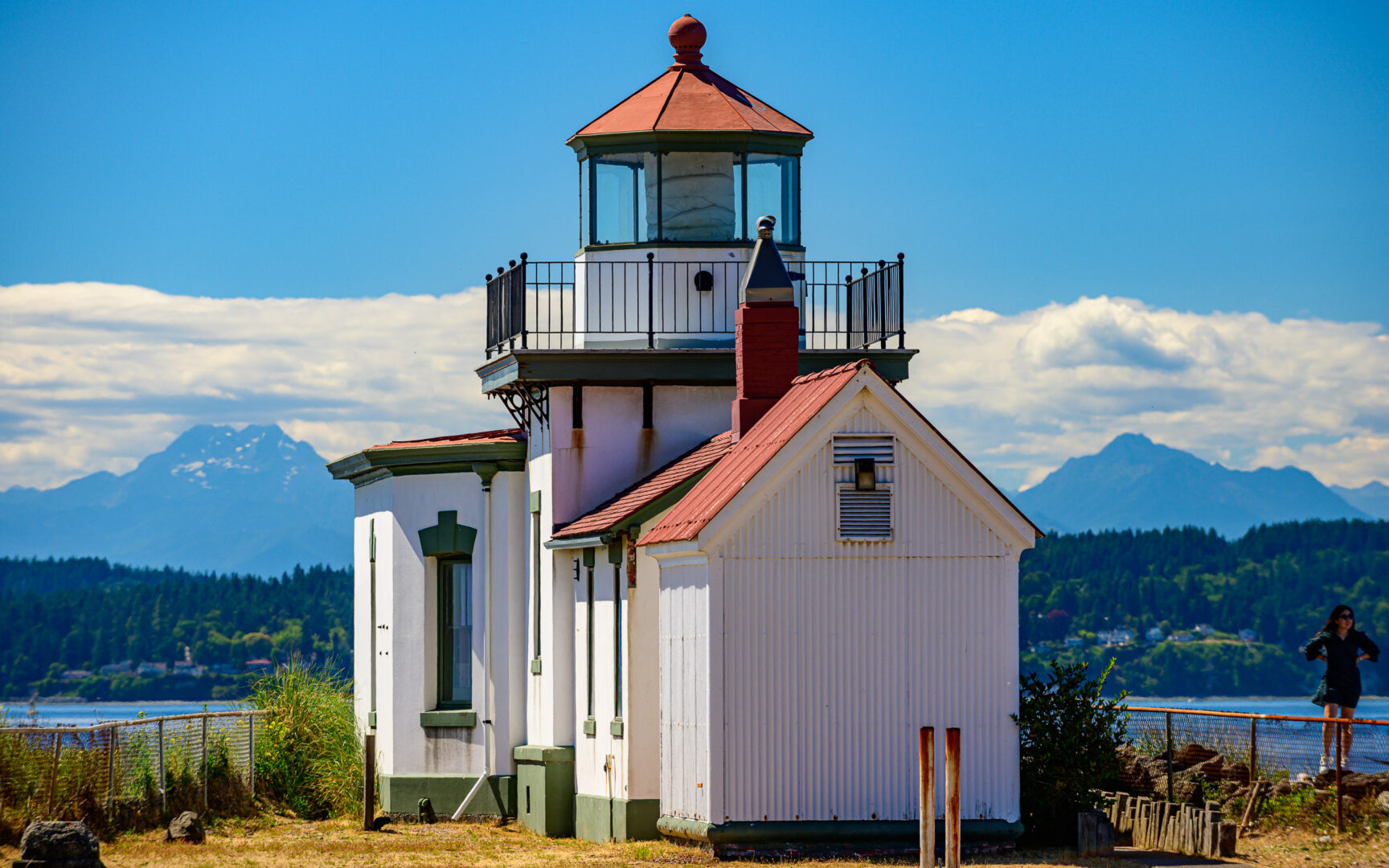 Color photo of the West Point Lighthouse at Discovery Park in Seattle. It is a white building with red roof. In the background are the Olympic Mountains, blue with distance, and capped with puffy white clouds.