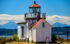 Color photo of the West Point Lighthouse at Discovery Park in Seattle. It is a white building with red roof. In the background are the Olympic Mountains, blue with distance, and capped with puffy white clouds.