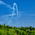 Color photo. Contrails lead from four Blue Angels in a tight formation, diving downwards. The sky is full of fading contrails from prior maneuvers. A green, tree covered slope is at the bottom of the frame.