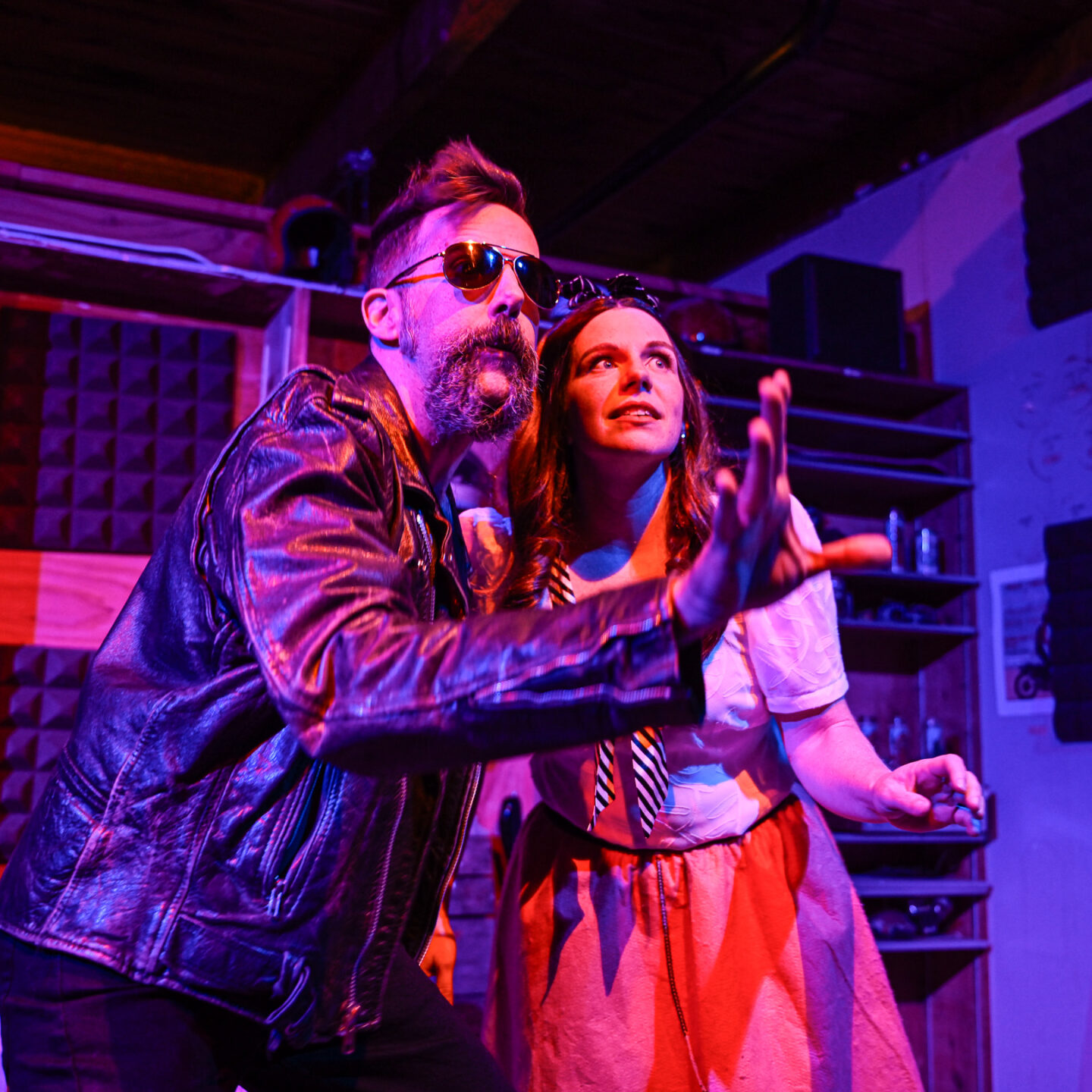 Photo of a man and woman, standing close, looking to the right. The man, on the left, is dressed in a black leather jacket and wears dark aviator sunglasses. His right hand is stretched out, fingers up, palm out. He has a short pompador hairstyle, and a fuzzy circle beard. The woman, on the right, has long hair, and is dressed in a white blouse and pink skirt. Her left hand is extended out, palm down.