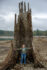 A person stands in the middle of a hollow stump on the shore of a rocky lake. Their arms are spread wide, and only barely touch the sides of the stump. The stump rises up to a jagged top, more than three times the height of the person standing in it.