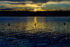 Color photo. Sunset. A body of dark water occupies the bottom two thirds of the frame. Small ripples reflect the dark blue of the sky at the edges of the frame, but the center is dominated by golden reflections of the setting sun, obscured above by dark clouds, and just about set behind a dark line of trees on the opposite shore. The remains of three pylons jut up out of the water in a line from left to right. The middle one is in silhouette, backlit by a column of light reflected on the water, pointing up at the hidden sun.