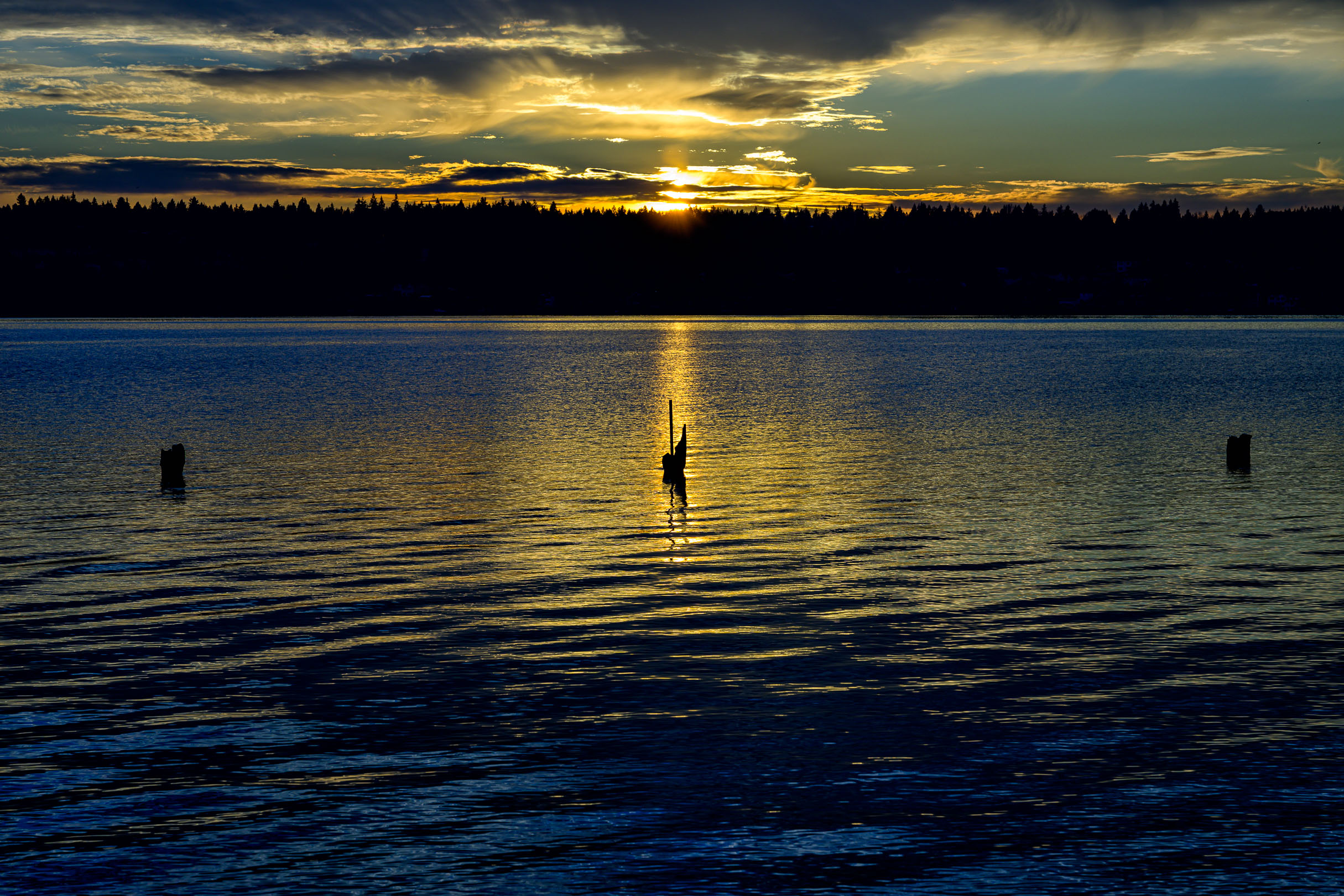 Color photo. Sunset. A body of dark water occupies the bottom two thirds of the frame. Small ripples reflect the dark blue of the sky at the edges of the frame, but the center is dominated by golden reflections of the setting sun, obscured above by dark clouds, and just about set behind a dark line of trees on the opposite shore. The remains of three pylons jut up out of the water in a line from left to right. The middle one is in silhouette, backlit by a column of light reflected on the water, pointing up at the hidden sun.