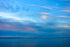 Color photo, in mostly blue tones. Sunset. High wispy clouds are pink from the unseen setting sun. Lower clouds are already mostly dark, but on the left, just above the distant mountains, sunset-pink shines through. A line thin line of clouds are below even that, separating the peaks from the hills below. In the foreground are the evening-dark waters of Puget Sound.