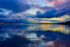 Color photo. Sunset. The waters of Lake Washington are in the foreground, covered in soft ripples, reflecting the active sky above. The sky is full of dark clouds, in shades of blue and purple, tinged pink in the upper right from the unseen setting sun. The line of the lake's far shore divides the photograph in two, between water and sky. The distant skyline of Seattle pokes up above the trees, into the clouds.