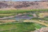 Color photo. A stream through a wetland, widening into a pond, surrounded by green and yellow grass. White birds are in the water. Arid, rocky hills of red stone rise up above the green grass.