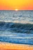 Color photo. The sun, barely risen above the blue ocean, in an orange sky, both blurred by distance. In the foreground, crossing the frame, is a wave beginning to crest, backlit by the sun and glowing a dark emerald. Below it the foam of a prior wave covers wet sand, which is reflecting the orange sky.