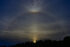 Photograph. The sun, just visible above a line of trees at the bottom of the frame. A faint halo, in pale rainbow colors, arcs completely around the sun, with a burst of color directly at the top of the arc. Wispy clouds are visible everywhere in the dark blue of the sky.