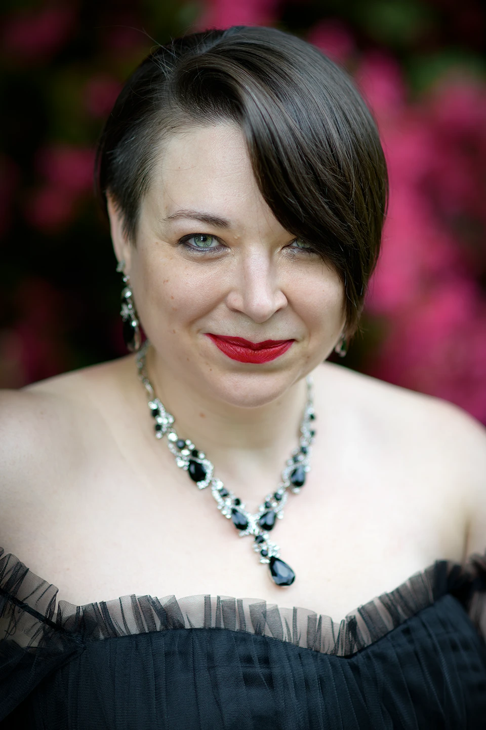 Color photo. A portrait of a woman in a black dress. She is wearing a silver neckalce with black stones. She has bright red lipstick and piercing blue eyes. She has dark hair, short on the right, with a long lock on the left almost covering her eye.