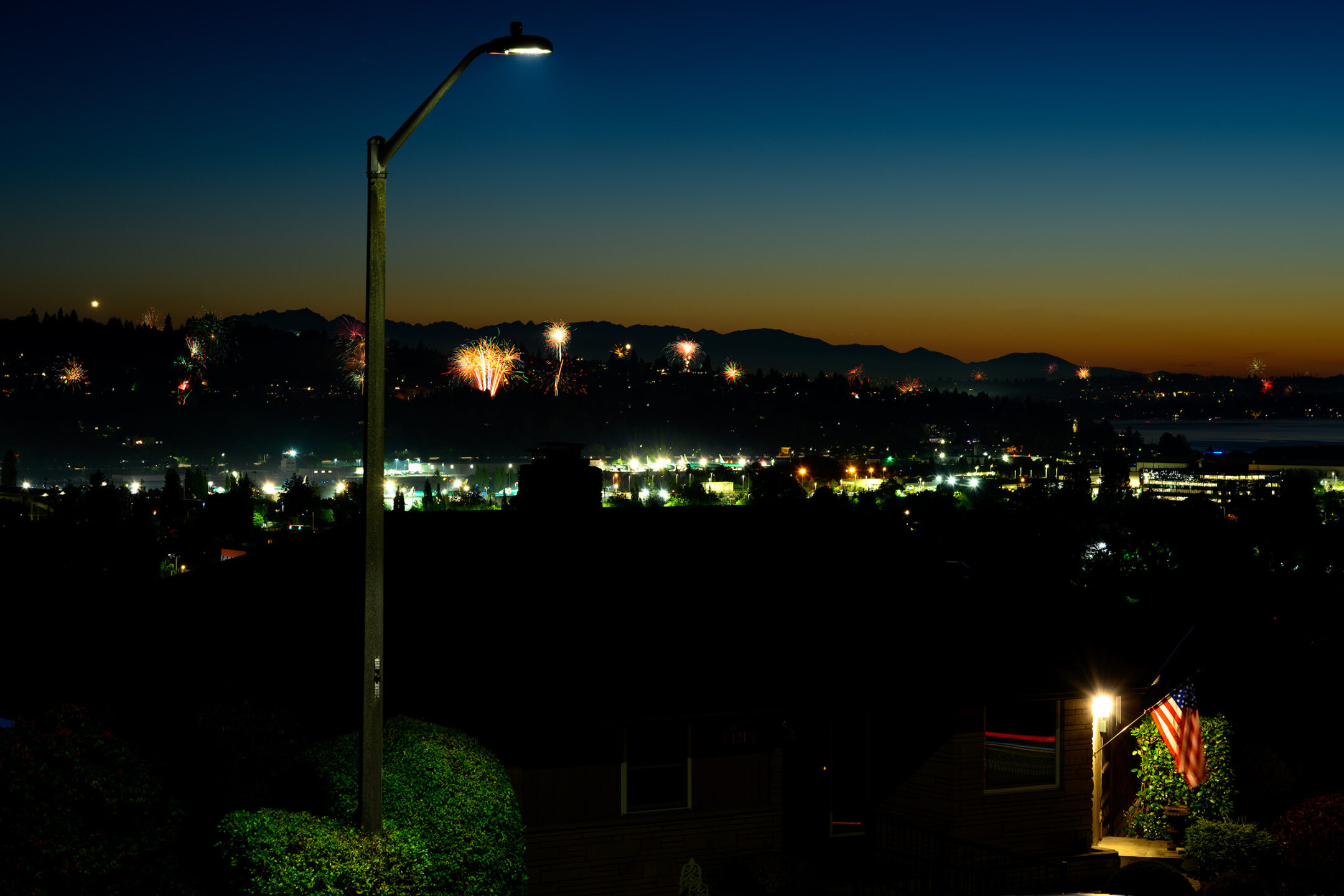 Color photo. A distant line of hills is visible against the dark red of a post-sunset sky. Fireworks are rising up everywhere from the hills. There are city lights dotting the hillsides. In the foreground is a streelight. In the lower left, an American flag hangs from a flagpole on the side of a house, lit by a floodlight.