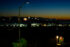 Color photo. A distant line of hills is visible against the dark red of a post-sunset sky. Fireworks are rising up everywhere from the hills. There are city lights dotting the hillsides. In the foreground is a streelight. In the lower left, an American flag hangs from a flagpole on the side of a house, lit by a floodlight.