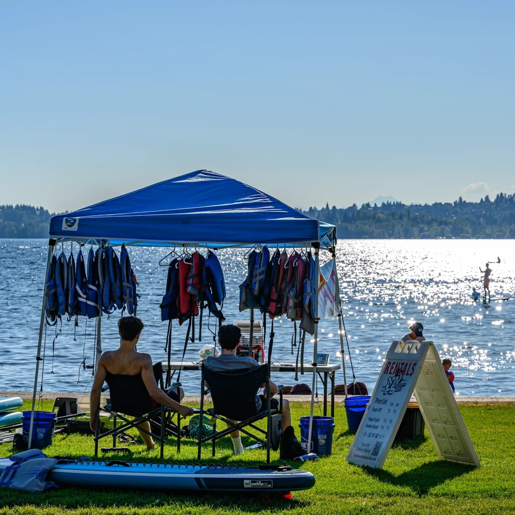 Color photo. A blue-roofed pop-up shelter, next to a lake. Two people are in its shade, sitting in front of a table, their backs to the camera, as they face the water. Life vests hang from the shelter above them, and a stand up paddle board rests on the grass behind them. Next to the shelter is a sign advertising rental rates for paddle boards.