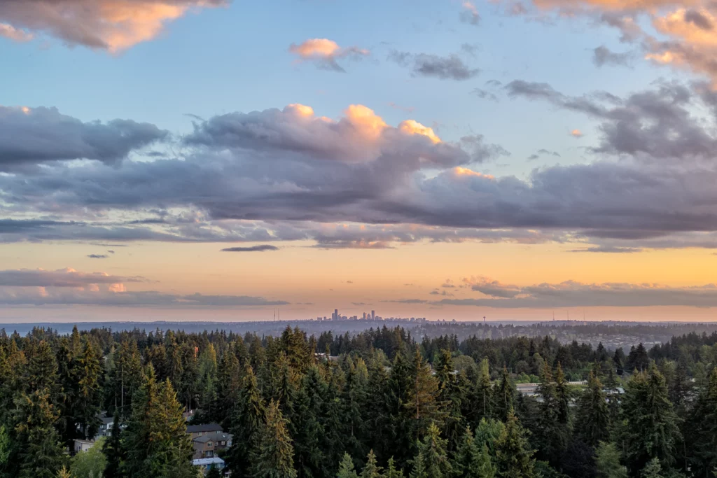 Color photo. The downtown Seattle skyline is seen in the far distance, beyond tree covered hills. The blue sky is filled with dark clouds edged in sunset orange.