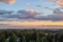 Color photo. The downtown Seattle skyline is seen in the far distance, beyond tree covered hills. The blue sky is filled with dark clouds edged in sunset orange.