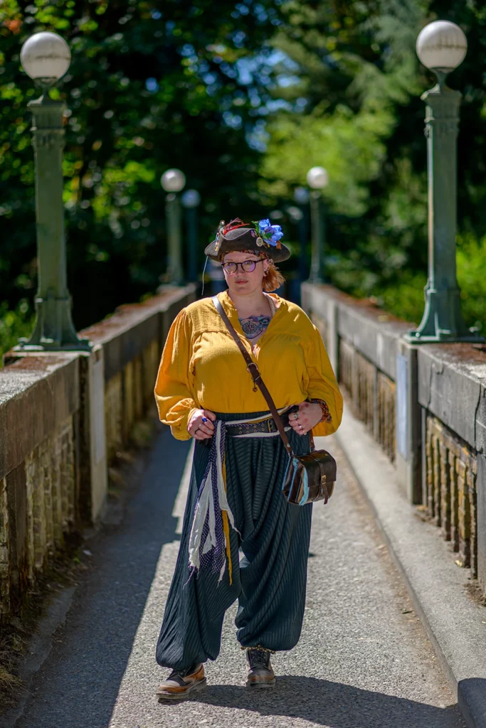 Color photo. A tattooed woman stands in the middle of a narrow bridge, lit by sunlight over her right shoulder, the concrete railings receding into the distance behind her. On the railings are a line of green light posts, topped with white globes. The woman is dressed as a pirate, in a honey yellow shirt and a tricorn hat covered in pins, with a large blue flower.