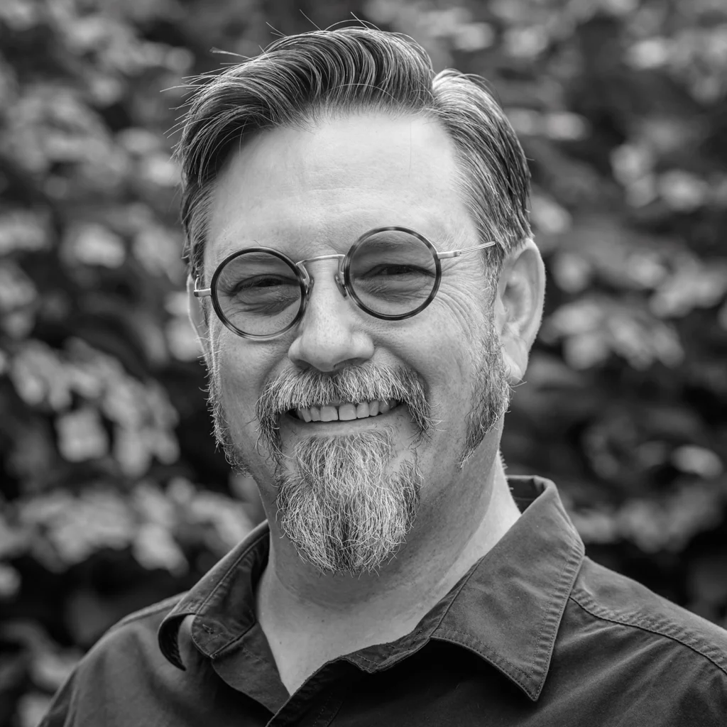 Black and white photograph. A smiling man, wearing round glasses. He has a goatee and muttonchop sideburns.