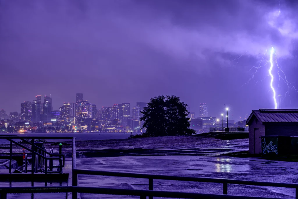 Color photo. The downtown Seattle skyline, seen from Gas Works Park on the north end of Lake Union. Night. The buildings are a patchwork of lit windows. The scene is lit purple by a lightning bolt on the far right of the frame, striking something just to the left of the Space Needle.