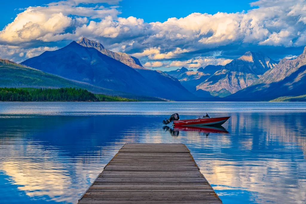 Color photo. Lake McDonald at Glacier National Park. A wooden dock extends from the bottom of the frame towards the center. Just beyond the dock a red speedboat is moored, 'Crestliner' written on its side. The gentle waves of the waters reflect the distant mountains, which are capped by clouds.