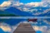 Color photo. Lake McDonald at Glacier National Park. A wooden dock extends from the bottom of the frame towards the center. Just beyond the dock a red speedboat is moored, 'Crestliner' written on its side. The gentle waves of the waters reflect the distant mountains, which are capped by clouds.