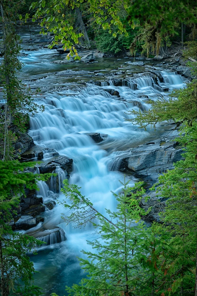 Color photo. A stepped waterfall, framed by trees, flowing over gray rocks. A long exposure has rendered the water as smooth, milky sheets.