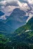 Color photo. A forested valley with steep sides leads up from the bottom of the frame towards a mountain, its jagged rocky sides rising up out of the tree line. Above, the sky is full of dark clouds.