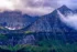 Color photo. Mount Cannon in Glacier National Park rises up on the right, its pyramidal rocky peak rising up out of clouds. On the left another mountain is visible, its peak obscured by dark clouds. Below that mountain are snowfield above forested slopes. Emerging from those slopes is a tall, thin waterfall, known as Bird Woman Falls.