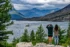 Color photo. A view of Saint Mary Lake, and Wild Goose Island, in Glacier National Park. Two people are standing on a rock in the foreground. The person on the left is wearing shorts and a green jacket, and is holding binoculars to their face. On their right is a person with long wavy dark hair, wearing a yellow flannel shirt and jeans. In the distance are rocky mountains below dark dramatic clouds.