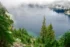 Color photo. Snow Lake, in Washington State, seen from a ridge above it. Tree covered slopes lead down to the water, which is a deep teal in the shallows. Clouds obscure the far shore.