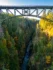 Color photo. An arcing bridge truss crosses the top of the frame. Below is the river gorge the bridge spans. The steep slopes are covered in evergreens, and dotted with autumn-yellow trees.