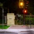 Color photo. Night. A deer walks from behind a metal box on a sidewalk, looking at the viewer. A street signal, showing yellow, and a crosswalk showing a red hand, are next to the deer. A fence behind the deer crosses the frame from left to right.