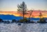 Color photo. Sunset. A lake, with a line of rocks sticking up from the water in mid frame. In the middle, from the largest pile of rocks, a pine tree grows. In the distance are mountains in blue shadow on the far side of the lake. Above, red and orange clouds catch the sunlight from behind the mountains.