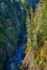 Color photo. A view of a river in a gorge. The left slopes are in shadow, and the right are in sunlight. Both are mostly covered in evergreens, with splashes of autumn yellow.