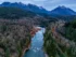 Color photo. A river extends up from the bottom of the frame, turning a corner to the right at mid-frame. On either side are bare trees, the hills above them dark with evergreens. At the top of the frame is a line of snow-capped mountains.