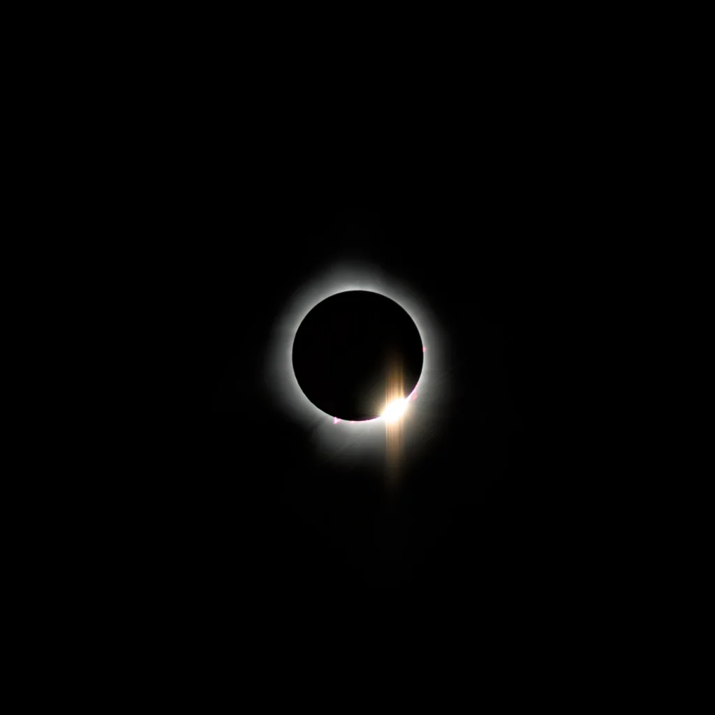 Color photo. The sun at full eclipse in 2024. The dark circle of the moon is surrounded by wispy silver corona, and purple solar prominences are visible. In the lower right, the sun peaks through mountains on the moon, it's brilliant yellow light spilling forth.