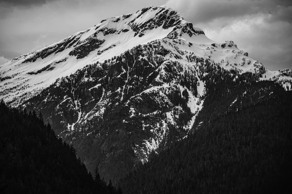 Black and white photo. A snow covered mountain, Elephant Butte, with dark trees and rock peaking through the snow. Its peak is mirrored below by two dark tree covered hillsides intersecting at the bottom of the frame.