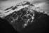 Black and white photo. A snow covered mountain, Elephant Butte, with dark trees and rock peaking through the snow. Its peak is mirrored below by two dark tree covered hillsides intersecting at the bottom of the frame.