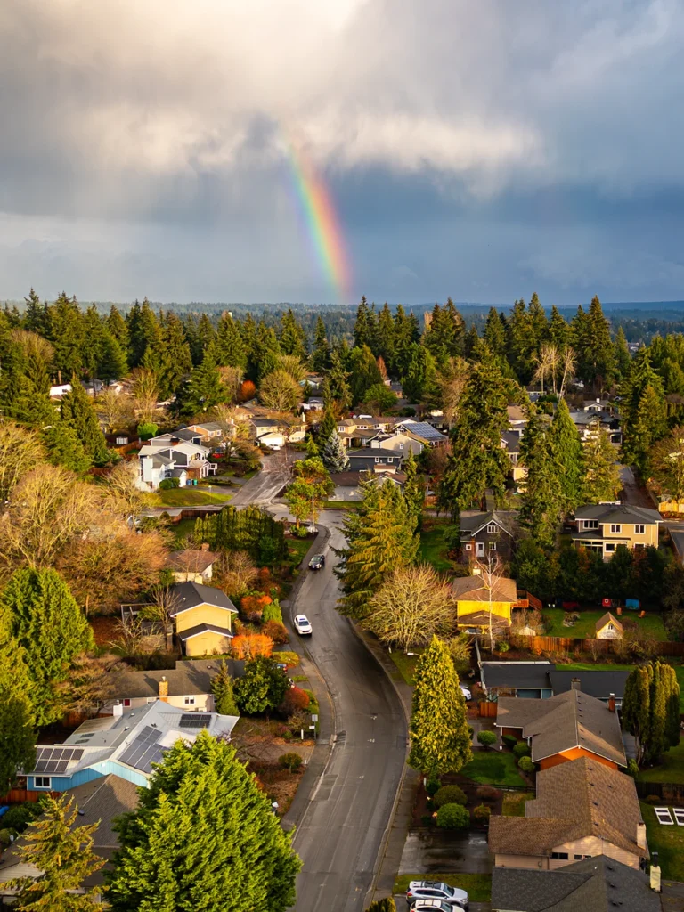 Color photo. A street, seen from above, winds up through a neighborhood from the bottom of the frame. Tall pines grow between the houses. The sky is full of dark clouds, but the foremost are lit by the sun from out of frame. A brilliant rainbow leads down from the clouds to the houses.