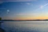 Color photo. A lake, reflecting golden sunset skies. The tree and house lined far shore is visible. On the left, the tiny silhouettes of people are seen, standing on the shore of the lake. Above them, in the distance, the peak of Mount Rainier is visible, catching the light of the setting sun, out of frame on the right. The sky above the horizon is golden, and shades to deep blue above, filled with wispy clouds. A prominent contrail leads in from the upper right to the center of the frame.