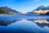 Color photo. Bowman Lake, in Glacier National Park, early morning. Mist rises from the still water, and clouds pool in the distant end of the valley, tall mountains on either side. The rising sun, out of frame, lights the clouds and the peak on the left. The mountains, sky, and clouds are reflected in the water at the bottom half of the photo.