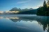 Color photo. Bowman Lake, in Glacier National Park, early morning. Tall pines are at the right of the photo, sunlight peaking through the branches. Mist rises from the still water. The tree-covered far shore recedes to the left, below shadowed mountain peaks. The water in the bottom half of the photo reflects the top half.