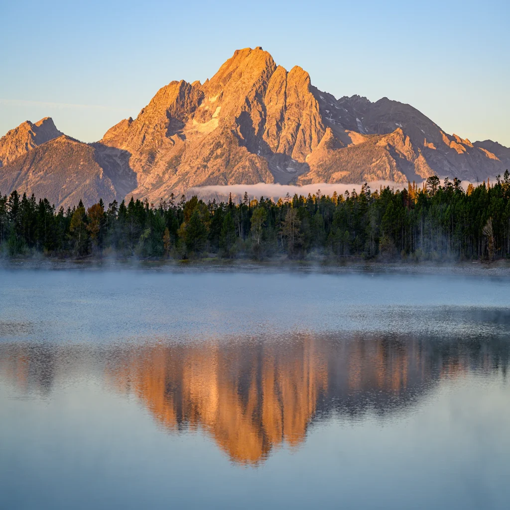 Color photo. Mount Moran, in Grand Teton National Park, lit by reddish early morning light. It is reflected in the misty waters of Colter Bay, on the far side of which is a tree lined shore.