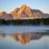 Color photo. Mount Moran, in Grand Teton National Park, lit by reddish early morning light. It is reflected in the misty waters of Colter Bay, on the far side of which is a tree lined shore.