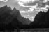 Black and white photo. Cascade Canyon, in Grand Teton National Park. The craggy peaks of the Cathedral Group are on the left, and the ridge along the canyon marches away from the viewer into the misty distance. In the foreground is a line of dark pines and a grassy slope. The sky is full of fluffy clouds, just covering the sun at the top of the frame, from which rays project onto the Cathedral Group.