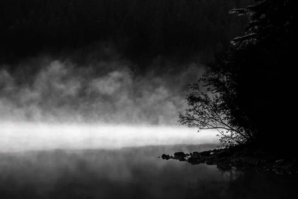 Black and white photograph. A line of brightly lit mist rises from the still waters of a lake. From the right, a dark tree covered promontory extends into the water, silhouetted against the mist. The tree covered far shore of the lake, beyond the mists, is just barely visible.