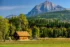 Color photo. A log cabin, golden in the morning light, in a field of grass, next to a line of trees beginning to turn color in the approaching autumn. Beyond the cabin a tree-covered hill rises, and beyond that a dramatic sheer peak, showing geological bands and strata. The blue sky contains wispy clouds.