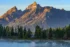 Color photo. Eagles Rest Peak, in Grand Teton National Park. The tall face of the mountain is lit by reddish morning light, with portions in deep shadow. At the bottom of the frame is a sliver of water below a tree lined shore.