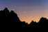 Color photo. The black silhouette of a jagged mountain range crosses the bottom of the photo. Above, the sky is a deep orange that fades to purple at the top of the frame. In the middle of the photo is the slender crescent of a waxing moon.