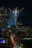 Color photo. The Space Needle, at night. Fireworks are seen launching from its roof, trailing sparks. A street leads from the bottom of the frame towards the Needle, full of cars.